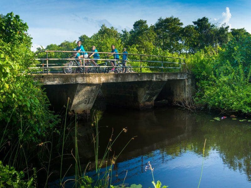 The photo views a group of cyclists ride on a river bridge